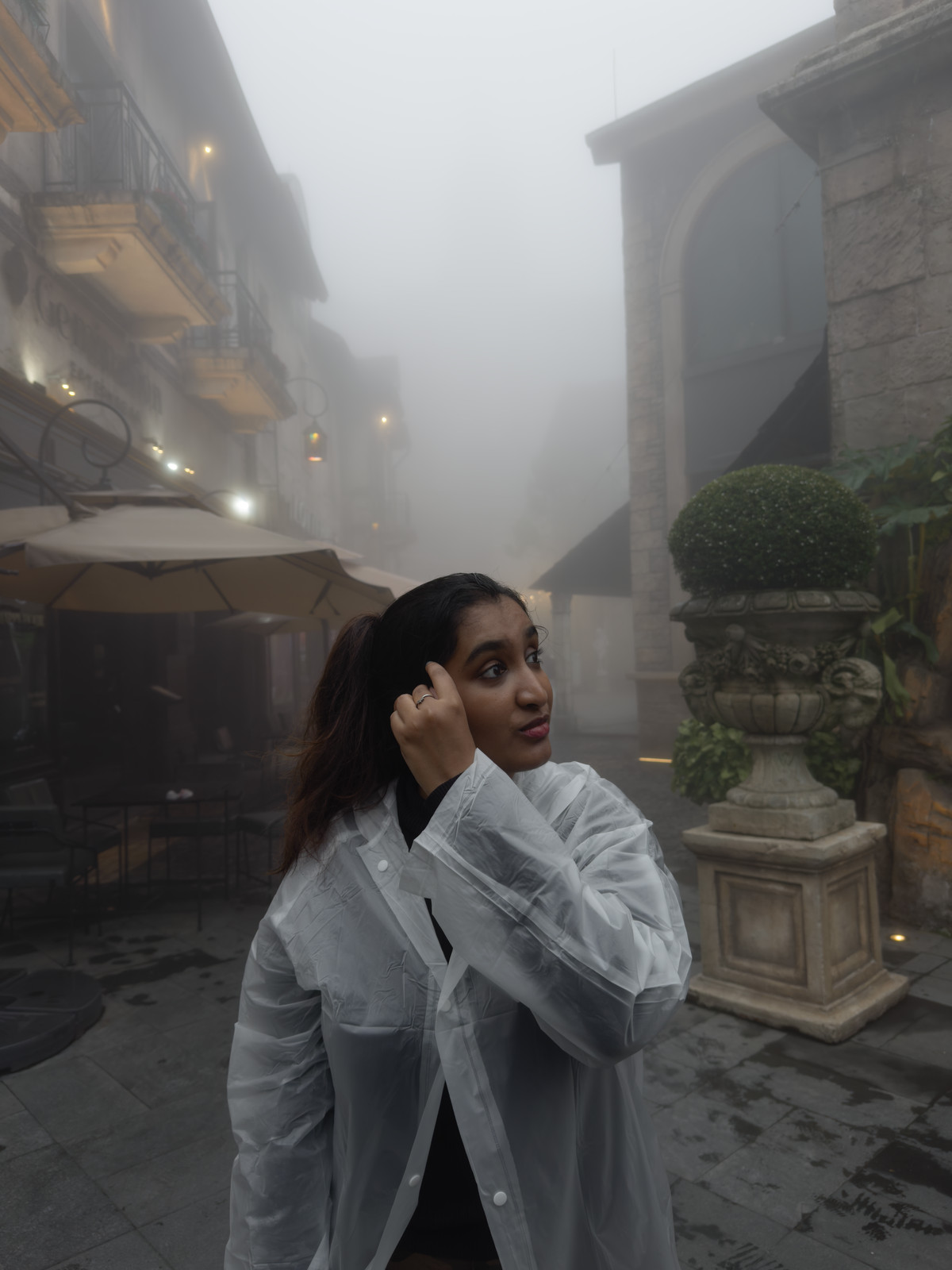 A woman in a white raincoat on a fog-filled street of old stone buildings, hand raised to her temple.