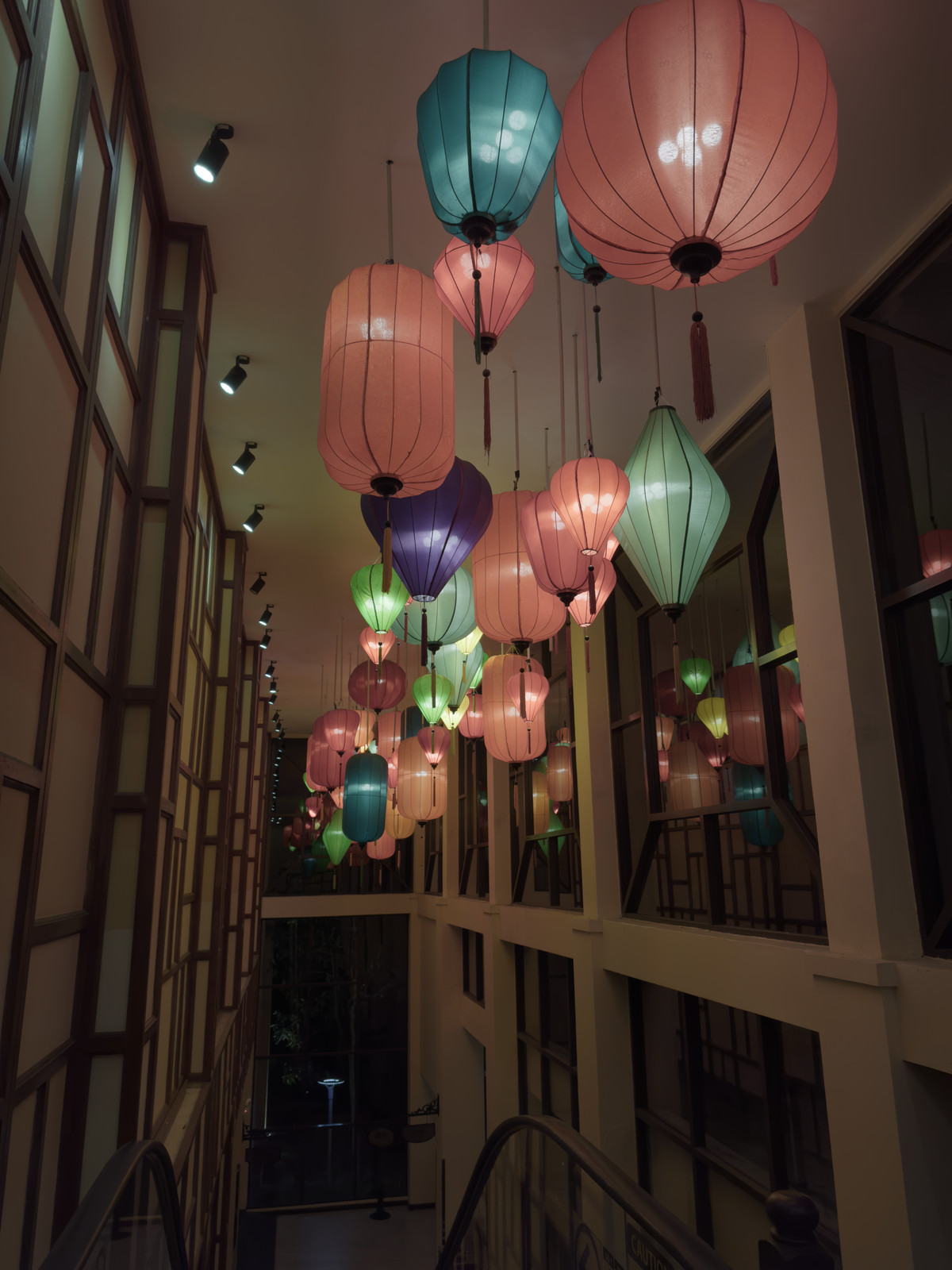 Silk lanterns in pink, teal and violet hanging down a tall wood-panelled stairwell.