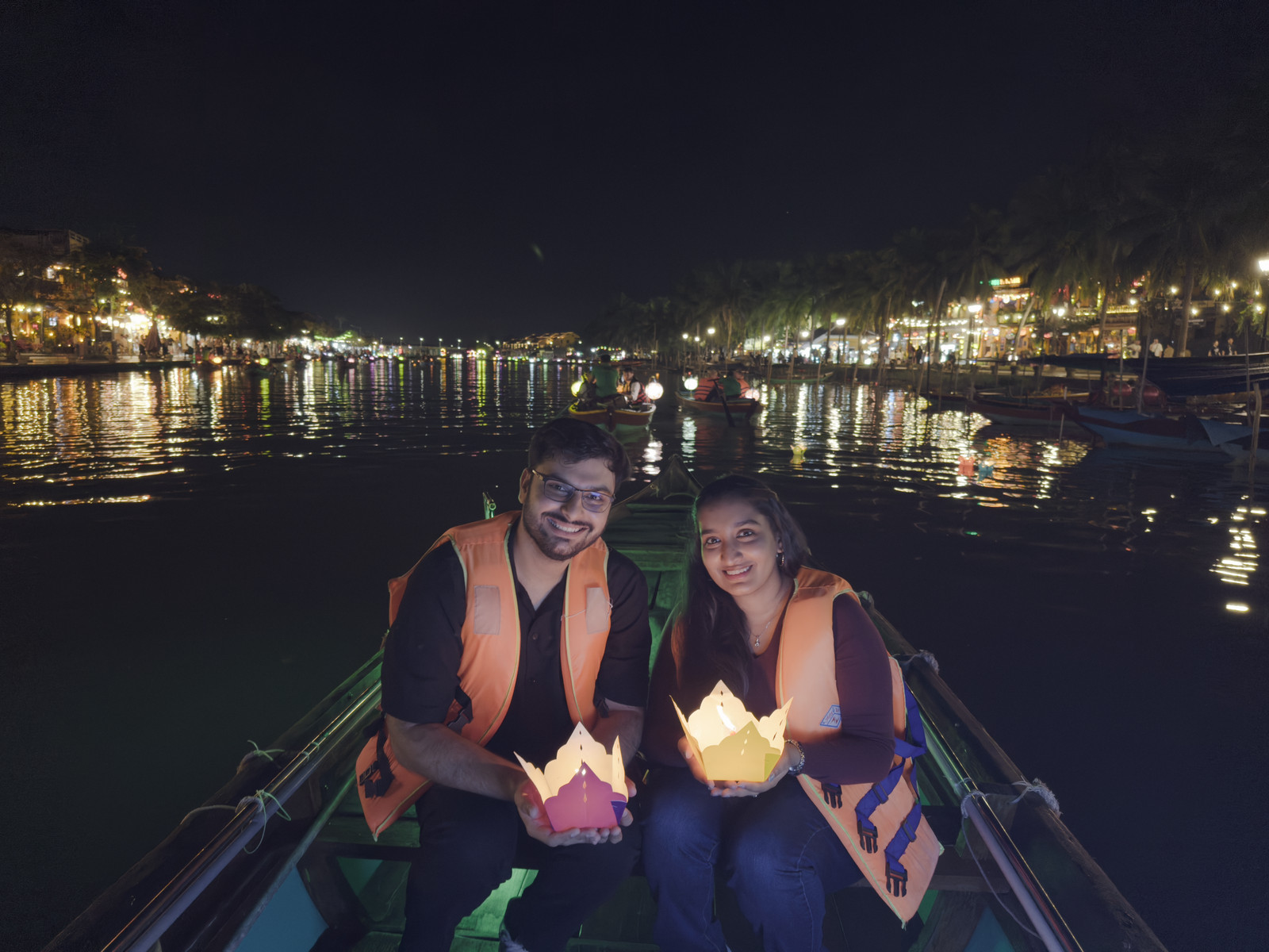 Two people in life jackets on a boat at night, each holding a lit paper lotus lantern, with a riverside town lit up behind them.