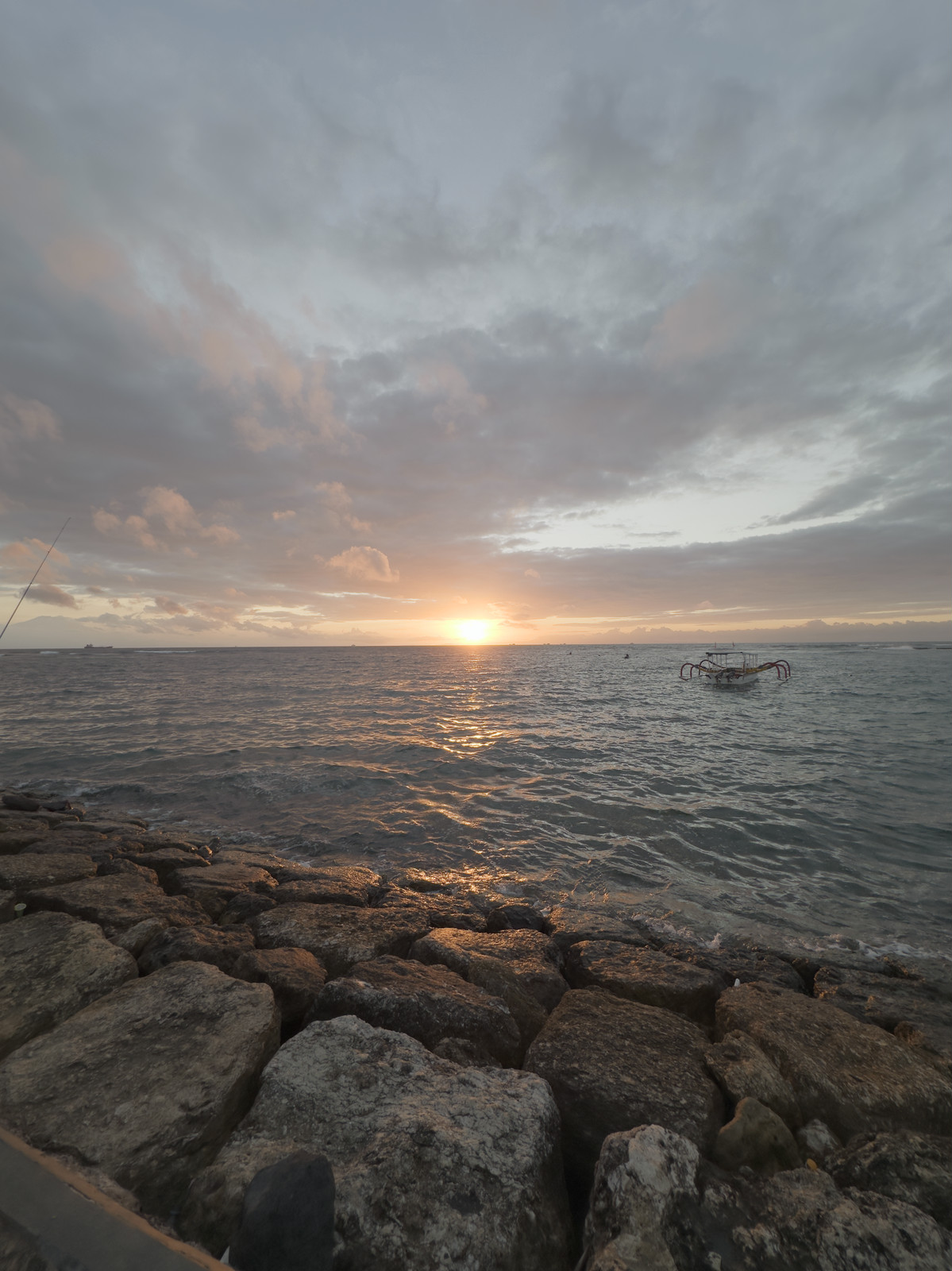 The sun setting over a calm sea, a rock breakwater in the foreground and a small outrigger boat offshore.